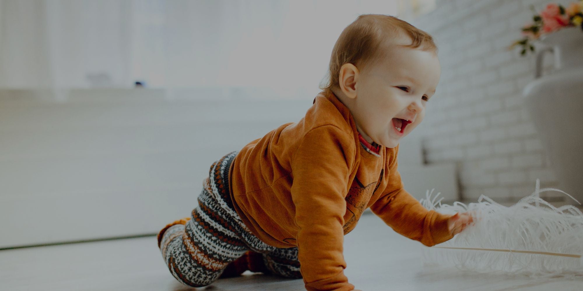 A baby crawling on a light floor in a bright room, wearing an orange long-sleeve top and patterned knit pants, reaching toward white fluffy decor.